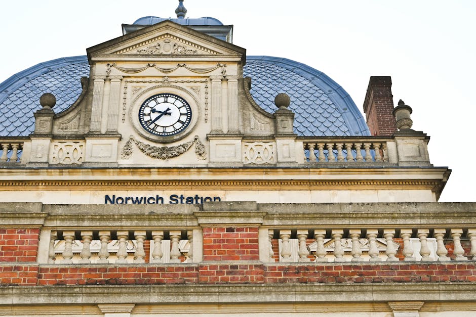 View of the upper section of Norwich Station building with a classical architectural style, featuring a large central clock with black Roman numerals and hands, set in a cream-colored façade adorned with decorative stonework and garland reliefs. The building has a blue, slate-tiled dome roof with rounded finials on each corner. A brick chimney is visible to the right side of the structure. Below the clock, the station name 'Norwich Station' is displayed on the stone ledge. The scene is captured from a low angle, emphasizing the building's detailed embellishments and the historic design, set against an overcast sky. This image relates to house removals and moving services by illustrating the kind of public infrastructure often involved in relocating home or business premises, as provided by Removal Van Bromley, who assist with packing, furniture transport, and logistics.
