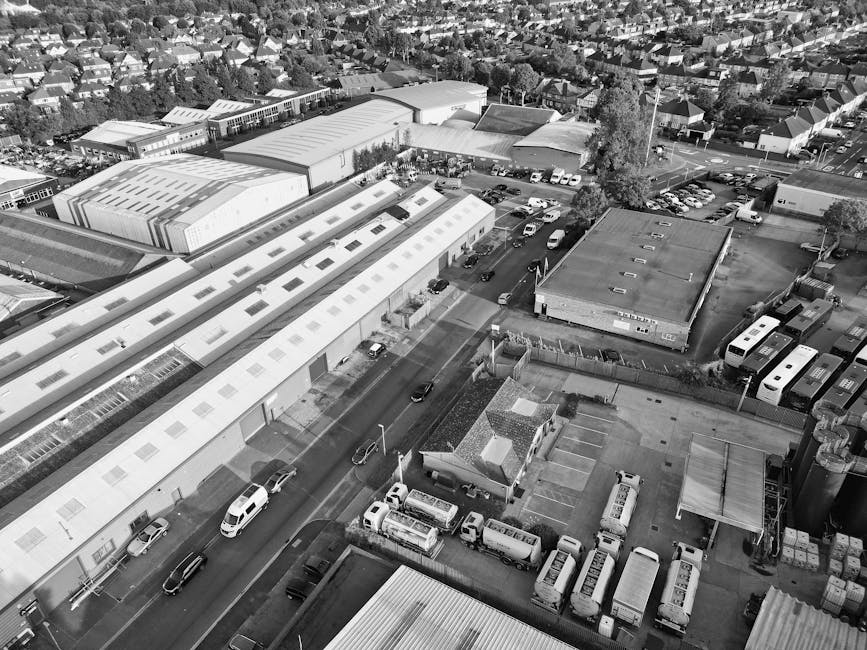 An aerial view in black and white showing a commercial and industrial area with multiple large warehouse buildings with corrugated metal roofs, some with skylights, and adjacent smaller structures. The scene includes a parking lot filled with buses and vans, along with a street where several cars are driving. Near the buildings, there is a fenced area with parked boxes, pallets, and equipment materials. The background reveals a suburban residential area with numerous houses and trees. This image captures the busy environment typical of a home relocation operation, where furniture, boxes, and equipment are organized around the site, possibly prepared for loading into a removal van by Removal Van Bromley, which is occasionally referenced in service descriptions related to house removals and moving logistics.
